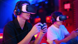 Two teen friends are playing a game console in VR headset and headphones using gamepads, talking and smiling while sitting on bean bags. Blue and red illumination virtual reality - Starpik