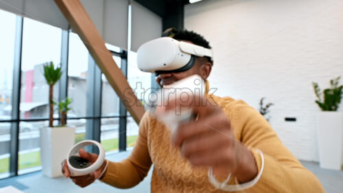 A black man exploring virtual reality using VR glasses and controllers in an office. Slow motion, virtual reality - Starpik Stock