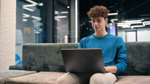 A young smiling man working using a laptop while sitting on a sofa in an office - Starpik