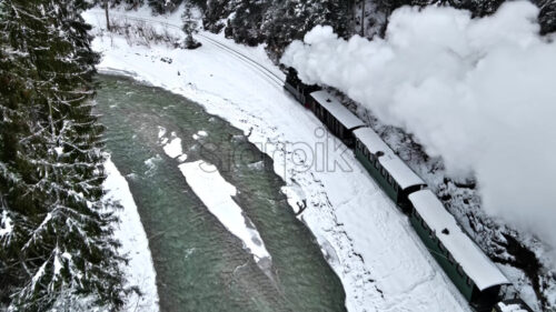 Aerial drone view of the moving steam train Mocanita in a valley along a river in winter, hills covered with bare forest and snow on mountains - Starpik