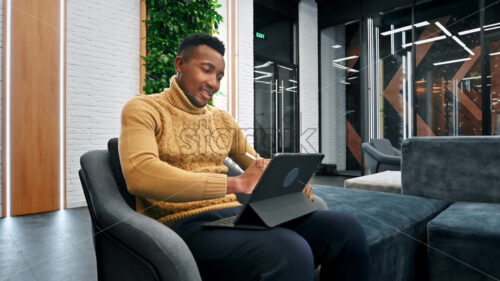 Black man working using a tablet while sitting on a sofa in an office. Slow motion - Starpik