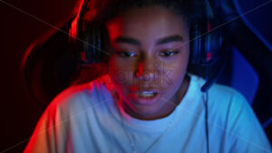 Black teen girl in headset playing video games in video game club with blue and red illumination, laughing and talking in voice chat. Slow motion - Starpik