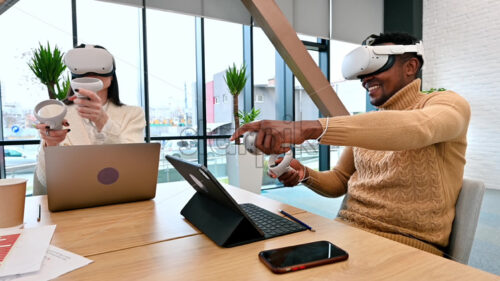 Business conference in VR in an office. A black man and caucasian woman using VR glasses and controllers, gadgets on the table. Slow motion, virtual reality - Starpik Stock
