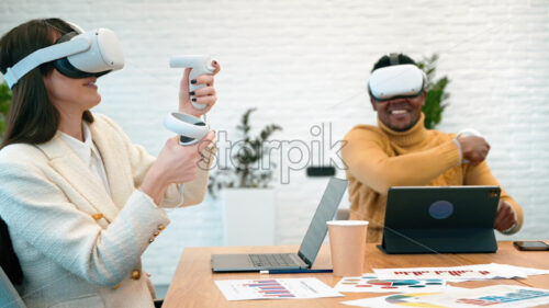 Business conference in VR in an office. A black man and caucasian woman using VR glasses and controllers, papers and gadgets on the table, virtual reality - Starpik Stock