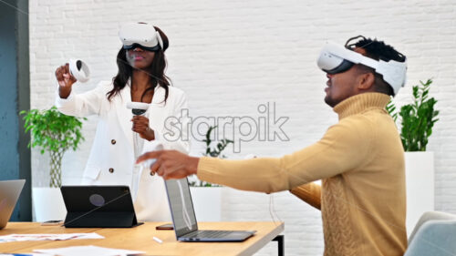 Business conference in VR in an office. A black man and woman using VR glasses and controllers, gadgets on the table. Slow motion, virtual reality - Starpik Stock