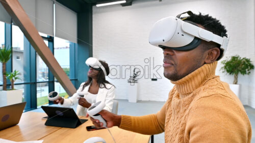 Business conference in VR in an office. A black man and woman using VR glasses and controllers, gadgets on the table. Slow motion, virtual reality - Starpik Stock