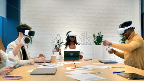 Business conference in VR in an office. Multiracial group of people using VR glasses and controllers, papers and gadgets on the table, virtual reality - Starpik Stock