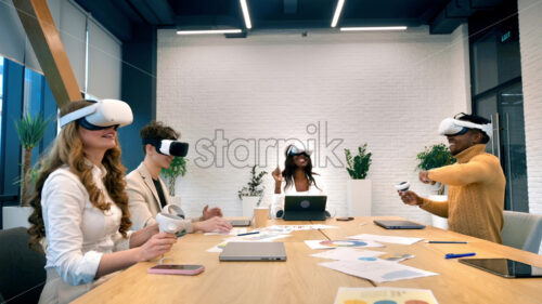 Business conference in VR in an office. Multiracial group of people using VR glasses and controllers, papers and gadgets on the table, virtual reality - Starpik Stock