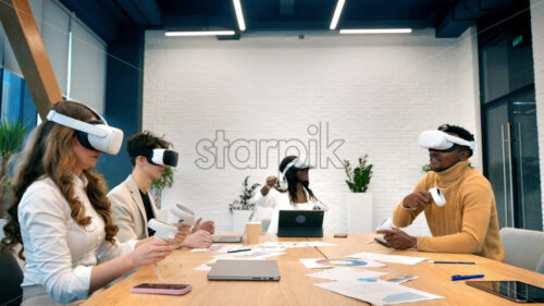 Business conference in VR in an office. Multiracial group of people using VR glasses and controllers, papers and gadgets on the table, virtual reality - Starpik Stock