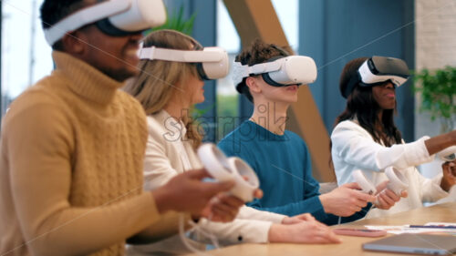 Business conference in VR in an office. Multiracial group of people. A young man teaching others how to use VR glasses and controllers, papers and gadgets on the table, virtual reality - Starpik Stock