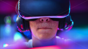 Close view of a teen playing a game console in VR headset and headphones using gamepad. Blue and red illumination, virtual reality - Starpik