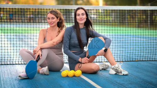 Two smiling women in sports suits posing with balls and rackets for playing Pickleball while sitting near the net on an outdoor court. Slow motion - Starpik
