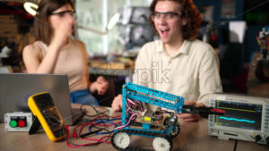 Two young happy engineers fixing a mechanical robot car in the workshop, computer programming - Starpik