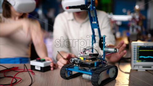 Two young happy engineers fixing a mechanical robot car in the workshop, using VR virtual reality headsets, computer programming - Starpik