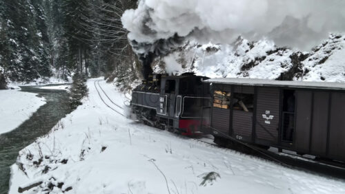 Video - Aerial drone view of the moving steam train Mocanita in a valley along a river in winter, hills covered with bare forest and snow on mountains