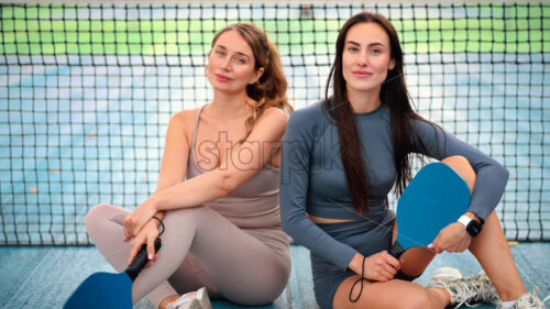 Video – Two smiling women in sports suits posing with balls and rackets for playing Pickleball while sitting near the net on an outdoor court. Slow motion - Starpik Stock