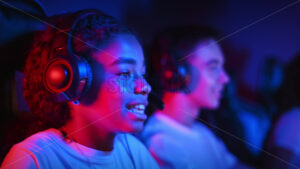 White boy and black girl teens in headsets playing video games in video game club with blue and red illumination, talking in voice chat. Slow motion - Starpik