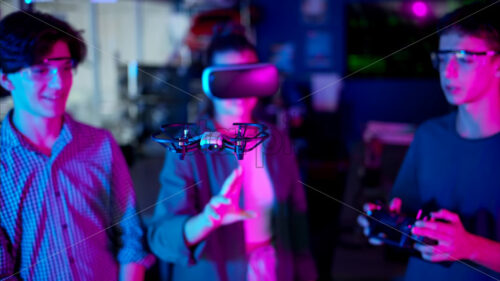 Young engineers with VR virtual reality headset controlling fliying drone in research laboratory, slow motion, neon blue pink lights - Starpik