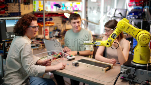 Young happy engineers programming an yellow robotic arm in the workshop to grab cardboard water glass, computer programming training for coffee preparation, celebrating success - Starpik