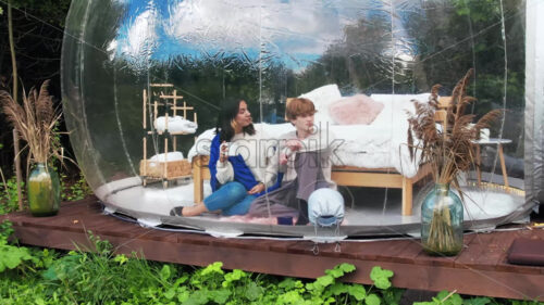 A couple sitting inside a transparent bubble tent at glamping. Sitting under a blanket, holding cups and talking - Starpik Stock