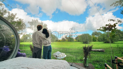 A couple sitting inside a transparent bubble tent at glamping. Sitting under a blanket, holding cups and talking - Starpik Stock