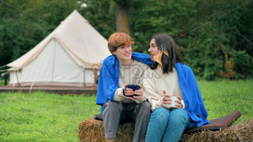 A couple sitting inside a transparent bubble tent at glamping. Sitting under a blanket, holding cups and talking - Starpik Stock