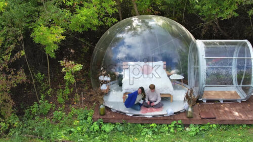 A couple sitting inside a transparent bubble tent at glamping. Sitting under a blanket, holding cups and talking, view from a drone. Lush forest on the background - Starpik Stock