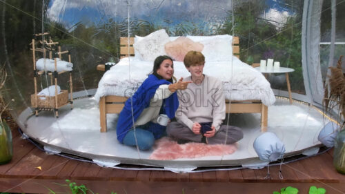 A couple sitting inside a transparent bubble tent at glamping. Sitting under a blanket, holding cups and talking. Lush forest on the background - Starpik Stock