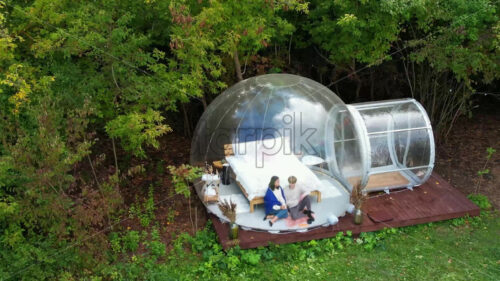 A couple sitting inside a transparent bubble tent at glamping. Sitting under a blanket, holding cups and talking. Lush forest on the background - Starpik Stock