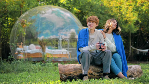 A couple sitting under a blanket on a stump, holding cups and talking at glamping. Tents on the background, lush forest around - Starpik Stock