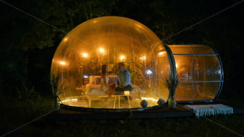 A couple talking while sitting inside transparent bubble tent with illumination at glamping at night. Lush forest on the background - Starpik Stock