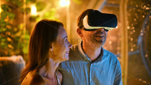 A hugging couple inside transparent bubble tent at glamping at night while man using VR glasses. Illumination inside - Starpik Stock
