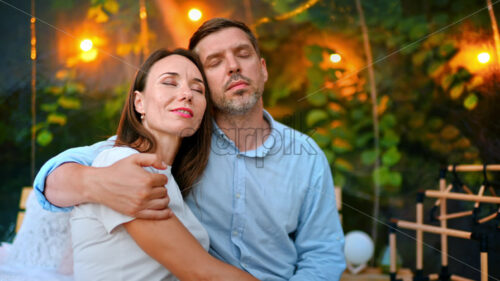 A hugging couple with closed eyes inside transparent bubble tent at glamping. Lush forest and illumination on the background - Starpik Stock