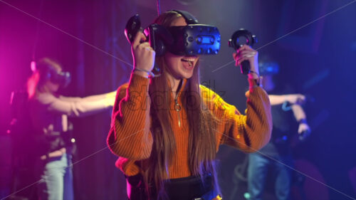 A joyful, interested girl is playing on VR treadmill with helmet and controllers. Other people playing nearby. Slow motion virtual reality - Starpik Stock