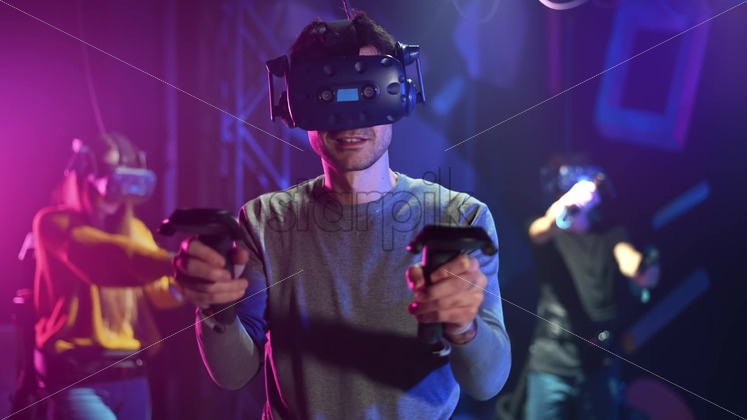 A joyful, interested man is playing on VR treadmill with helmet and controllers. Other people playing nearby, virtual reality - Starpik Stock