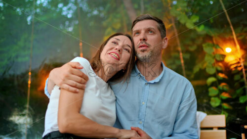 A talking and hugging couple inside transparent bubble tent at glamping. Lush forest and illumination on the background - Starpik Stock