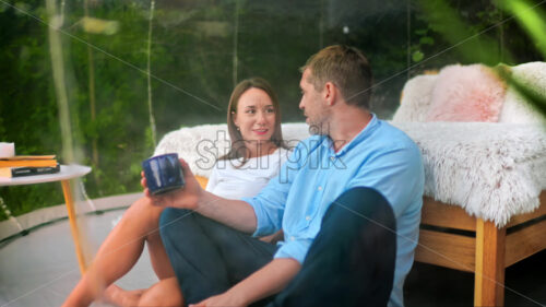 A talking couple sitting inside transparent bubble tent at glamping with cups. Lush forest on the background - Starpik Stock