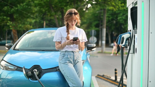 A young blonde woman using smartphone at a car charging station with charging electric car nearby. Chisinau, Moldova. Slow motion - Starpik