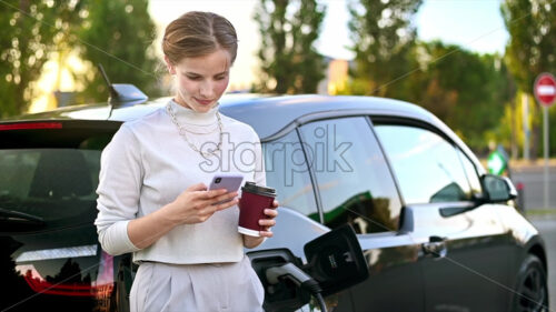 A young blonde woman with coffee using smartphone at a car charging station with electric car nearby in Chisinau at sunset, Moldova. Slow motion - Starpik