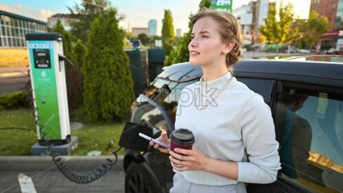 A young blonde woman with smartphone and coffee at a car charging station with charging electric car nearby in Chisinau, Moldova - Starpik