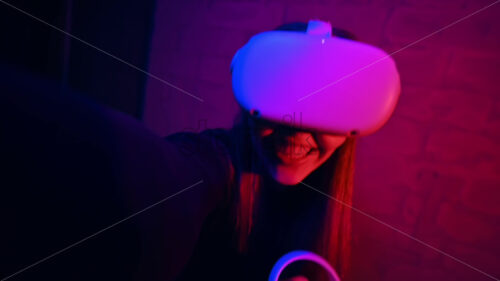 A young interested woman playing in VR games using VR glasses and controllers, filming herself on camera. Red and blue illumination. Slow motion - Starpik Stock
