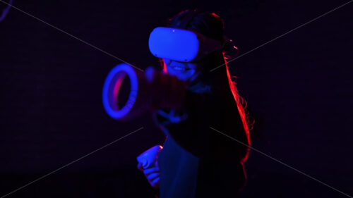 A young interested woman playing in VR games using VR glasses and controllers, red and blue illumination. Slow motion virtual reality - Starpik Stock