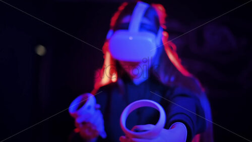 A young interested woman playing in VR games using VR glasses and controllers. Red and blue illumination. Slow motion virtual reality - Starpik Stock