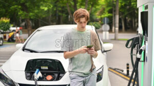 A young man using smartphone at a car charging station with charging electric car nearby. Chisinau, Moldova. Slow motion - Starpik Stock