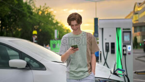 A young man using smartphone at a car charging station with charging electric car nearby. Chisinau at sunset, Moldova. Slow motion - Starpik Stock