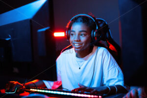 Black teen smiling girl in headset playing video games in video game club with blue and red illumination. Keyboard with illumination - Starpik Stock