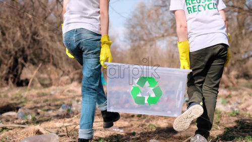 Boy and girl in rubber gloves holding a container at plastic garbage collection in a polluted clearing, recycling signs on the T-shirts. Slow motion - Starpik