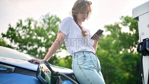 CHISINAU, MOLDOVA – JULY, 2022: A young blonde woman using smartphone at a car charging station with charging electric Renault ZOE. Slow motion - Starpik