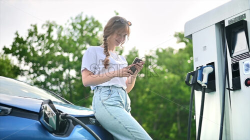 CHISINAU, MOLDOVA – JULY, 2022: A young blonde woman using smartphone at a car charging station with charging electric Renault ZOE. Slow motion - Starpik
