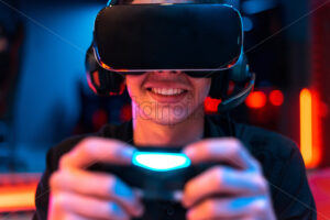 Close view of a smiling teen playing a game console in VR headset and headphones using gamepad. Blue and red illumination - Starpik Stock